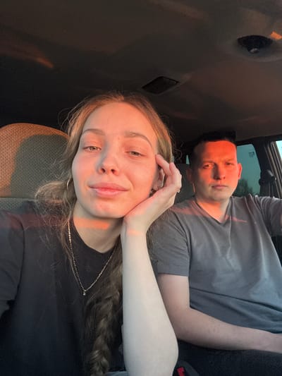 A selfie shows a young woman with long, wavy hair, wearing a black shirt and a silver necklace, leaning her head on her hand. Behind her is a man wearing a grey t-shirt. They appear to be inside a vehicle. The image is tilted. There is no information available to confirm the location is in Ivanovka, Russia.