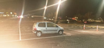A silver hatchback is parked awkwardly in an outdoor parking lot at night. The car straddles a parking line, and the parking lot is lit by bright overhead lights. Orange traffic cones line an adjacent area. Other vehicles are visible in the distance. A building is lit up in the background. The ground is asphalt, and parking lines are painted white.