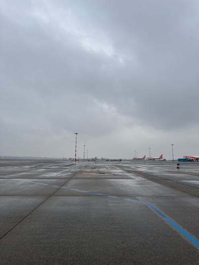 The image depicts an airport tarmac under an overcast, uniformly grey sky, indicating a dull, potentially wet day. The ground, a vast expanse of dark grey asphalt and concrete, is reflective with moisture, suggesting recent rain. A prominent blue curved line marks the foreground pavement, with additional white and faint red lines and rectangular panel divisions visible. In the mid-ground, a series of red and white striped poles extends diagonally into the distance. Several white ground support equipment vehicles are parked further back. The background features multiple commercial aircraft, predominantly orange-tailed and white-bodied, consistent with EasyJet livery. At least four such aircraft are identifiable, with a blue ground service vehicle positioned next to the nearest aircraft on the right. A low horizon line with indistinct distant shapes separates the tarmac from the sky. No individuals, specific actions, or interactions are visible. This scene is identified as Milan Malpensa Airport (MXP), situated in the city of Somma Lombardo, Italy.