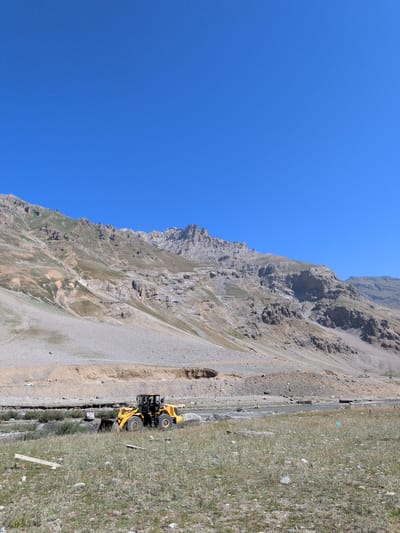 In the mountainous region of Matayeen, India, a yellow front-end loader is engaged in construction work near a rocky riverbed. The loader is positioned on the left side of the frame, its bucket lowered towards the ground. In the background, a vast, arid mountain range dominates the landscape, with sparse vegetation clinging to its slopes. The sky is a clear, cloudless blue. The scene suggests ongoing infrastructure development in a remote, natural setting.