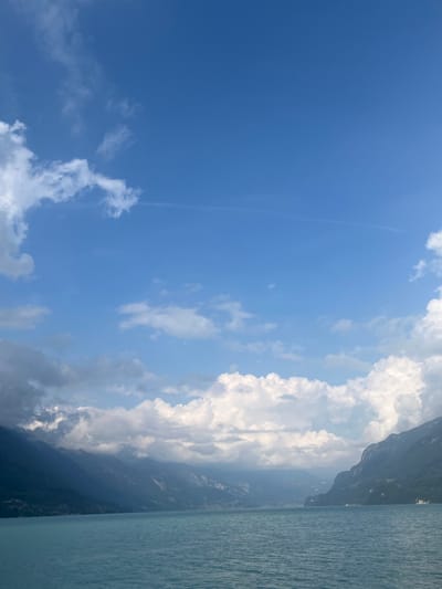 The image presents a wide, horizontal landscape view of a large body of water, consistent with Lake Brienz, located in the vicinity of Bönigen, Switzerland. The upper approximately two-thirds of the frame consists of a vibrant blue sky, featuring scattered white cumulus clouds. A faint, linear contrail is visible traversing the upper mid-sky. The lower third of the frame shows the calm, turquoise-blue surface of the lake, exhibiting minor, diffuse ripples. Steep, mountainous terrain, predominantly dark green to bluish-grey, flanks both sides of the lake, receding into the distant background. Portions of the mountain peaks are obscured by a mix of white and grey clouds. Distant structures, likely part of small settlements, are faintly visible along the base of the mountains where they meet the water. No individuals, vehicles, or specific human activities are discernibly present within the captured scene.