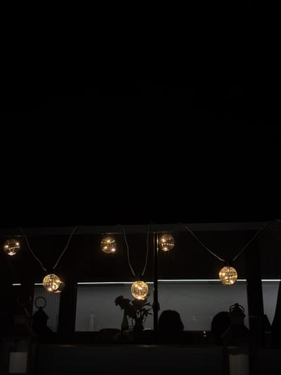 The image depicts a string of illuminated, globe-shaped fairy lights suspended across a dark background. These lights are positioned above a darkened interior space, partially illuminated by the glow of the lights. In the foreground, on the left, is a lantern with a white candle inside. To the right, a second lantern is visible, also containing a candle. Behind the string lights, a window reveals a dimly lit interior with the silhouettes of two people visible. A vase with some foliage is also present on a surface behind the lights. The overall scene suggests an evening setting, possibly a restaurant or a decorative outdoor space. The location is identified as the city of Grad Šibenik, Croatia.
