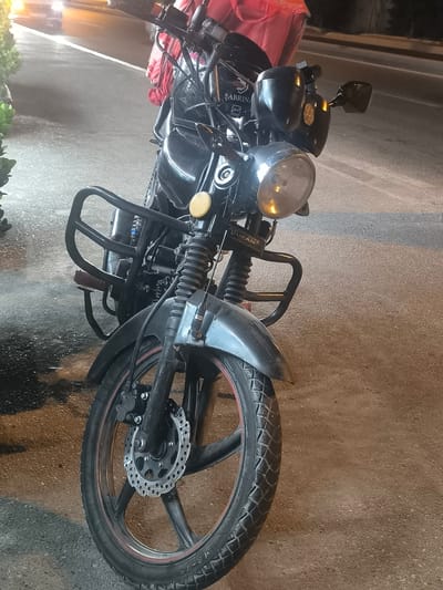 A black motorcycle is parked on an asphalt road at night. The motorcycle has a red delivery bag on the back. The motorcycle has a single headlight and two rearview mirrors. The front wheel has a disc brake. There is a metal crash bar in front of the engine. The background shows a street with a white line and some lights.