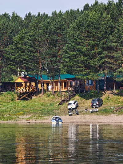 The image depicts a waterfront scene in Podporozh'e, Russia, featuring a wooded hillside with a few buildings and vehicles near the water's edge. A dense forest of tall pine trees forms the backdrop, extending across the top of the frame. On the slope, a cluster of wooden structures with yellow walls and green roofs are visible. One building has an elevated wooden porch with a red roof.
In front of the buildings, on a sandy and grassy incline, are two vehicles: a white SUV with a trailer and a black SUV with a trailer. A small white and blue motorboat is docked at the edge of the water in the foreground. The water itself is calm, reflecting the sky and the surrounding greenery. The overall atmosphere is that of a rural retreat or a riverside settlement.