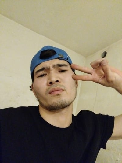 A young man, wearing a blue baseball cap and a black t-shirt, poses for a selfie. He has dark hair and a light beard. His right hand is raised to his face, with his index and middle fingers extended in a peace sign, positioned near his cheek. His expression is neutral. The background appears to be an indoor setting with a light-colored wall, possibly a room with plain walls and a ceiling. There is a circular vent or fixture visible on the wall in the upper right portion of the image. The lighting suggests it might be daytime or an indoor artificial light source is used. The overall mood of the image is casual and self-aware. There is no visible text in the image. The location context is Tuzdybastau, Kazakhstan.