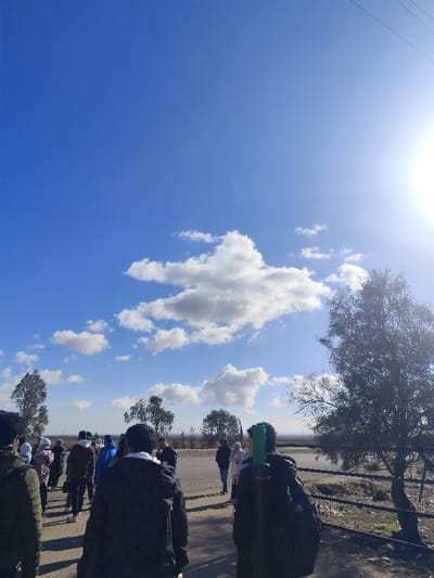 A group of people walks away from the viewer on a road. The sky is blue with scattered clouds. A few trees stand on the right, casting shadows. The sun shines brightly in the upper right corner, creating glare. The land beyond the road appears dry and slightly elevated.