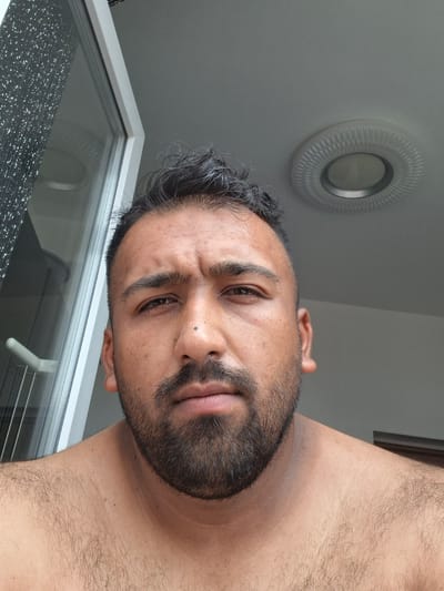 A man with dark hair and a beard, possibly of Middle Eastern or South Asian descent, is looking directly at the camera. He has visible sweat on his forehead and is shirtless, showing a hairy chest and shoulders. The background includes a window with a patterned texture on the glass and a circular light fixture on the ceiling. The man is positioned in the foreground, taking up most of the frame. The image appears to have been taken indoors, possibly in a residential setting, with a glimpse of an outdoor view through the window. The lighting suggests it is daytime. The man's facial expression is neutral. The scene does not provide enough context to determine the specific location within Evere, Belgium, other than that it is an interior shot with elements that could be found in many urban dwellings.
