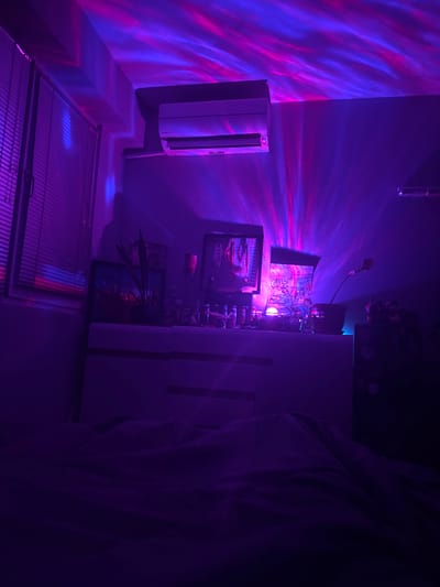 The image presents an interior scene, possibly a room, characterized by a vivid, predominantly purple and blue lighting scheme. The light projects patterns onto the walls, ceiling, and floor, creating an abstract, almost dreamlike ambiance.
Prominently visible are several objects, including what appears to be a wall-mounted air conditioning unit, various pieces of artwork or pictures hanging on the wall, shelves, and some decorative objects or plants. The floor is visible in the lower portion of the frame.
The presence of a person is suggested on the right side of the image, but only a portion of their clothing is visible, rendering their identity and actions unclear.
There is no clear indication that the location is in the city of Novi Pazar, Bulgaria.