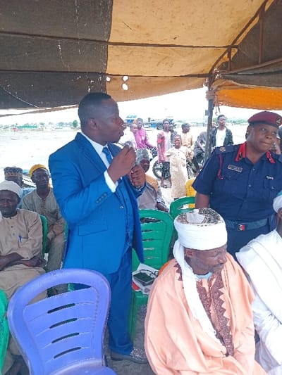 A man in a blue suit is speaking into a microphone in front of a group of people sitting in plastic chairs under a white tent. The man in the suit is standing with his left hand on his hip. He is addressing the seated people, including a man in a white robe and a female officer in a dark blue uniform with a badge on her chest. There are several other individuals standing and sitting around the seated group, some of them appear to be listening to the speaker. The scene is likely taking place at an outdoor event in Katabu, Nigeria. The background shows a grassy area with a road behind the tent. There is also a motorbike in the background, which is partially visible. The location is close to the Niger River. There are trees and vegetation in the background. The overall mood is formal and engaging.