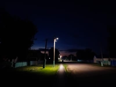 The image depicts a nighttime scene on a paved road with a sidewalk on the left. A single streetlamp illuminates the sidewalk and the edge of the road. On the left side of the sidewalk, there is a grassy area with a fence and trees in the background. On the right side of the road, there are houses and more fences. The sky is dark blue, indicating it is either late evening or early morning. The scene appears to be a quiet residential area, possibly a village or small town, with no people or vehicles visible. The location is identified as Qobda, Kazakhstan.