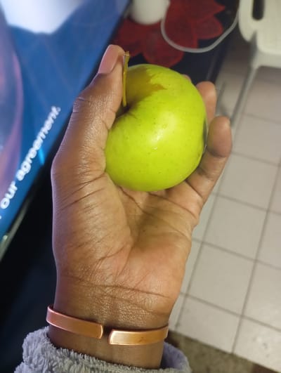 A close-up, slightly overhead shot details a human hand holding a green apple within an indoor domestic setting in Johannesburg, South Africa. The hand, exhibiting a dark skin tone, securely grasps the round, light-green apple, which shows small water droplets on its surface and retains a short, brown stem. A metallic, copper-toned cuff bracelet is worn on the wrist, and a portion of a grey, textured sleeve is visible at the bottom of the frame. The background to the upper left features a blurred electronic screen displaying white text including "our agreement" against a dark blue field. To the upper right, a tiled floor with light-colored square tiles and darker grout lines is visible. Further in the background, out-of-focus elements include indistinct red shapes, a white object, and a thin white cord.