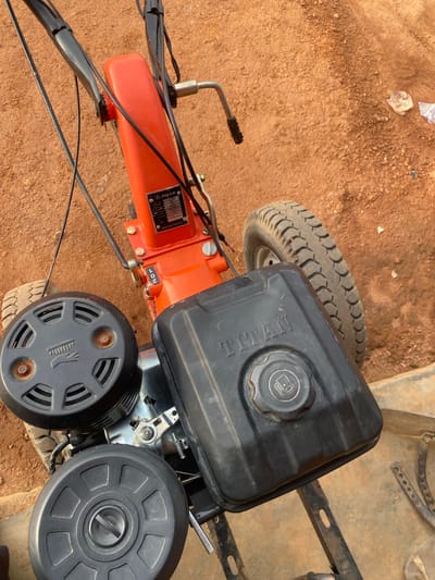 This is a close-up, high-angle shot of an orange and black agricultural tiller, likely a power tiller or walking tractor, resting on reddish-brown dirt ground in Jere South, Nigeria. The tiller is stationary. The foreground is dominated by two black circular components, one above the other, with ridged patterns. Above these, a black fuel tank with the embossed word "TITAN" is visible. To the right of the fuel tank, a black tire with a visible tread pattern is partially in frame. The tiller has an orange body with black handles extending upwards and out of frame. A metal label with text is affixed to the orange body. The surrounding environment appears to be an outdoor setting with loose soil and some discarded plastic items in the background. The lighting suggests it is daytime.