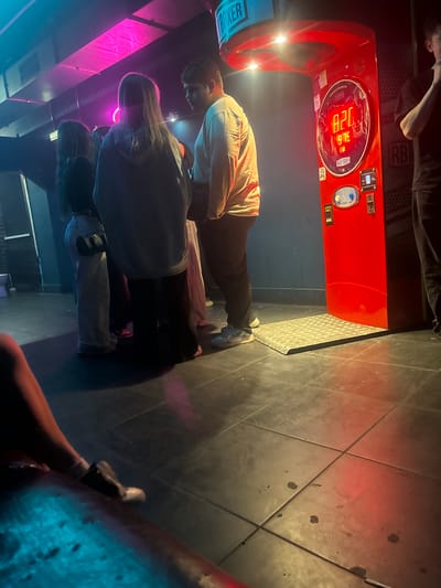 The image depicts a dimly lit indoor scene, likely a bar or entertainment venue, with multiple individuals present. A prominent red punching bag arcade game, labeled "ROCKY BOXER," is visible, displaying a score of "916" and "426". The game appears to be in use or has recently been used, as people are gathered around it. One person, wearing a light-colored shirt and dark pants, is standing near the game, possibly having just played or about to play. Another group of people are seen in the foreground, with one individual in a grey shirt and dark pants, and another in a light-colored top and dark pants. A third person is partially visible, wearing a grey hoodie and light-colored pants. The floor is tiled, with some appearing to be scuffed or dirty, reflecting the ambient lighting. The scene is illuminated by a combination of the arcade game's lights, neon pink and blue lighting from a nearby structure, and overhead lights. The overall mood appears casual and social, suggesting a night out. The time of day cannot be determined, and weather conditions are irrelevant as it is an indoor setting. There is no indication of the specific location beyond the "ROCKY BOXER" branding on the arcade game.