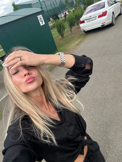 A woman with blonde hair is lying on the ground, holding her hand to her forehead. She is wearing a black shirt and a watch on her left wrist. Her eyes are closed, and her lips are slightly parted. A white car is parked in the background, and a green building with the number "6" on it is also visible. The scene appears to be outdoors, possibly in a park or recreational area. The weather seems to be cloudy. The text "E007EA 193" is visible on the license plate of the car. The overall mood of the image is one of relaxation or perhaps weariness.