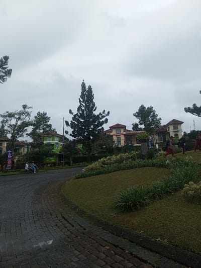 This image depicts an overcast daytime scene in a residential area of Sukanagalih, Indonesia. The sky is a uniform light gray, indicating a cloudy or overcast day.
In the foreground, a curved, paved road made of dark bricks or stones sweeps from the lower left towards the mid-left of the frame. To the right of the road, there's a well-maintained grassy slope adorned with various shrubs and smaller plants, suggesting landscaped grounds.
The midground and background are dominated by several large, multi-story houses, each featuring light-colored facades, possibly stucco, and distinctive reddish-brown tiled roofs. One house on the left side of the road stands out with its bright green exterior. Tall, slender pine trees are prominent features, especially one in the center, alongside other leafy trees that contribute to a lush, green environment around the residences.
Several people are visible. On the left side of the road, two individuals are seated on a motorcycle, seemingly stopped or moving slowly. Further to the right, on the landscaped grassy area near the houses, three people are walking. They appear to be wearing red shirts or tops with dark bottoms, and one individual seems to be carrying a bag or backpack, possibly indicating staff or a specific group.
A blue and red banner or flag is partially visible on the left side of the road, near the houses. Visible text on the banner includes "NEW" and "10%". The overall scene suggests a quiet, upscale community with well-maintained properties and landscaping, under a muted sky.