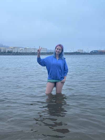 A young woman with dyed blue and pink hair stands in the calm water of a harbor, likely in Gijón, Spain, as suggested by the presence of industrial structures and storage tanks in the background, typical of a port city. She is wearing a blue zip-up hoodie and green shorts with white polka dots. Her right hand is raised, forming a peace sign. The sky is overcast and the water is choppy.