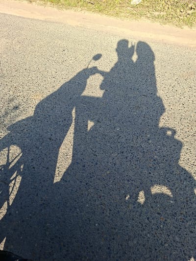 The image shows the shadow of a person on a motorcycle cast on an asphalt road. The shadow is elongated and distorted, indicating a low sun angle. The road is bordered by grass on the left side. The wheel of the motorcycle is visible at the bottom right of the image. The exact location in Uleeblang, Indonesia cannot be determined from the image alone.