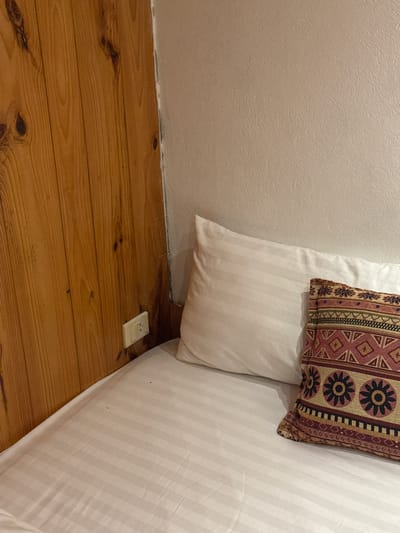 The image shows a corner of a room. A white wall is visible on the left side. The floor is made of wooden planks. An electrical outlet is located near the wall. A bed with white striped sheets and pillows is located on the right side of the image. A pillow with a geometric pattern sits atop the white pillow. There are no people visible in the image. This image was taken in Du Già Commune, Vietnam.