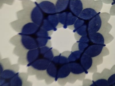 The image shows a pattern of stylized floral elements arranged in circles. The "petals" of each flower are blue, with short lines extending outward from the centers of the intersections. The backgrounds around the flowers are white, with a soft, muted gray outlining the floral shapes, creating a blurred, shadow-like effect. The overall impression is a repeated, stylized floral design on a surface.