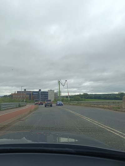 The image shows a view from a vehicle traveling on a road in Cambridge, United Kingdom. The road appears to be a dual carriageway with two lanes of traffic. In the foreground, the dashboard and windscreen of the vehicle are visible, reflecting the scene ahead.
On the left side of the road, there is a red cycle lane separated from the main road by a metal barrier. Beyond the barrier, there is a sidewalk and then a modern building with a facade of brown and grey panels, and visible concrete and scaffolding on the right side of the building. This appears to be a construction site. A tall, green construction crane with a red and white section is visible on the right side of the image, also suggesting ongoing construction.
Several cars are on the road: a dark SUV in the right lane, a dark-colored car in front of it, and a blue car in the left lane. A red car is also visible further ahead in the left lane. In the distance, there are green fields and trees under a cloudy sky. The overall atmosphere is overcast and possibly damp, judging by the reflections on the road. A single bird is seen flying in the sky on the left side of the image.