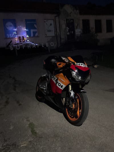 The image shows a motorcycle parked in front of a building at night. The building appears to be an old structure with posters and decorative lights on its facade. The text "ВОРНАТЕВО" is visible on the building, indicating the location is Bornarevo. The motorcycle is orange and black, with "REPSOL" branding. A Michelin Man sticker is visible on the front fender. The ground is asphalt, and there are some scattered leaves and debris. The overall scene suggests a nighttime street view in a rural or semi-urban setting.