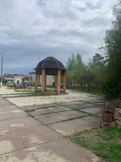 The image depicts a park scene in Kremyonki, Russia. A brick gazebo with a dark, weathered roof is centrally located, positioned on a paved walkway made of large concrete slabs with grass growing in between the joints. In the background, a playground with colorful equipment is visible, along with several multi-story buildings. The sky is overcast with a power line running across the upper-left corner of the image. A metal object, possibly a rusty barbecue, sits on the paved area near a grassy patch. The vegetation consists of trees with light green leaves.