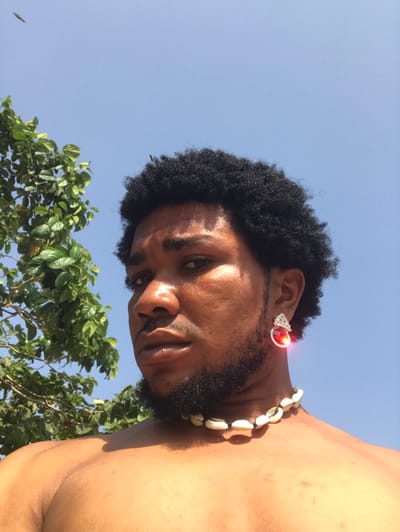 An outdoor, upward-angle portrait depicts an adult male in the city of Igbologun, Nigeria, under a clear blue sky. The subject, a male with a dark complexion, black afro-textured hair, and a beard, looks directly at the viewer. He is shirtless, revealing his upper torso and shoulders. He wears a choker-style necklace made of white cowrie shells. A prominent earring is visible in his right earlobe, featuring a silver-toned setting surrounding a large, oval, reddish-orange gemstone that reflects bright light. A portion of a green-leafed tree is present in the upper left background. Strong sunlight illuminates the scene, casting highlights on the subject's skin and the earring.