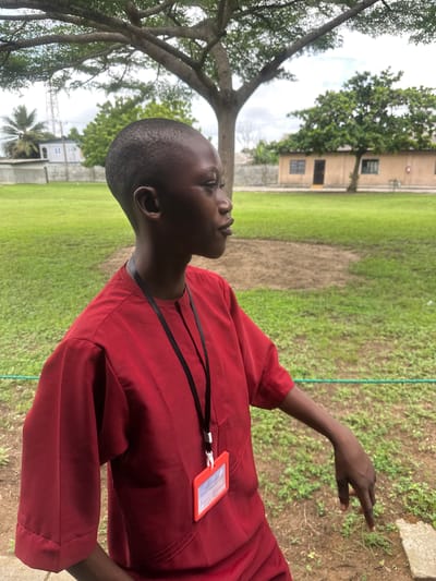 A young person with dark skin and a short haircut is shown from the chest up, facing to the right. They are wearing a red tunic with short sleeves and a black lanyard with an orange ID badge attached. The background is an outdoor scene with a large tree overhead, a grassy field, and a light brown building with windows and a door. The building appears to be a single-story structure with a corrugated roof. In the distance, there are other trees and a palm tree. A cell tower is visible on the left side of the image. The lighting suggests it is daytime, possibly overcast. The overall setting is rural or suburban.