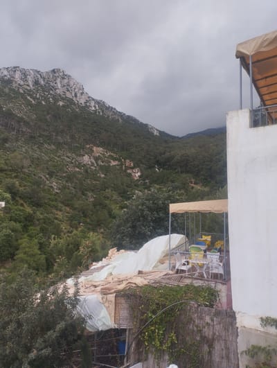 This image presents a scenic view of a mountainous landscape, likely near Tétouan, Morocco, under an overcast sky.

In the foreground on the right, a white building wall with a flat roof structure is partially visible. An upper balcony or terrace, shaded by a beige canopy and featuring a metal railing, extends from the building. Below it, a lower patio area is equipped with a similar beige canopy, providing shade over several colorful plastic chairs (white, yellow, green, and blue) and a white plastic table, suggesting an outdoor dining or relaxation space.

To the left and slightly below this patio, a rustic structure is covered with various light-colored tarpaulins and canvas sheets, possibly serving as a storage area or a makeshift greenhouse. A wooden slat fence, heavily draped in green vines, runs across the lower part of the foreground.

The midground and background are dominated by a majestic mountain range. The closest mountain face features rugged grey rock formations interspersed with dense clusters of pine trees and other evergreen vegetation. Further into the background, the mountains are entirely covered in lush green forests, extending up to the horizon.

The sky is uniformly grey and cloudy, indicating an overcast day. No human activity is explicitly visible, but the arranged furniture on the patio suggests a space prepared for occupants. There is no visible text in the image. The overall scene conveys a sense of natural beauty and tranquility, characteristic of mountainous regions like those surrounding Tétouan.