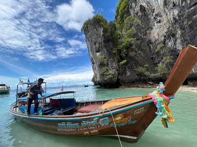 This media file depicts a man standing on a longtail boat in the turquoise waters of Ton Sai, Thailand. The boat, adorned with colorful flower garlands at its bow, is moored near a dramatic limestone karst island covered in lush green vegetation. The water is calm and shallow, revealing the sandy seabed in places.

The man, wearing a blue and white shirt with the word "THUNDER" visible and dark pants, appears to be a boat operator or guide. He is standing on the edge of the boat, looking towards the interior. Several life vests are visible on the boat, suggesting it is prepared for passengers.

In the background, other boats and a larger ferry with passengers can be seen on the horizon. The sky is a vibrant blue with fluffy white clouds, indicating fair weather. The bright sunlight suggests it is daytime, likely late morning or early afternoon. The scene conveys a sense of tropical tranquility and adventure.

Visible text on the boat includes Thai script and the words "6752 01237", "PHI PHI ISLAND VILLAGE", and "PariPat Tour", indicating the boat's affiliation and possibly its registration number.