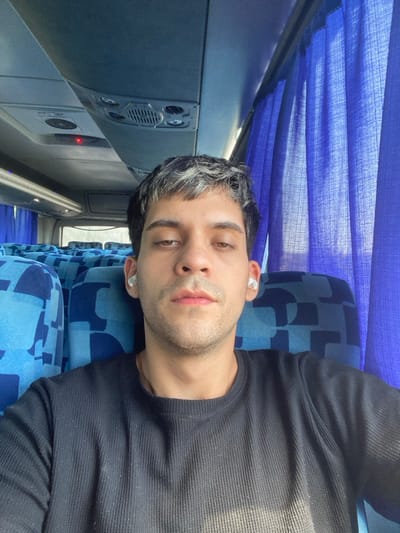 The scene depicts the interior of a passenger bus, reportedly located in Belén de Escobar, Argentina. An adult male, appearing to be in his 20s or 30s, is captured in a selfie-style shot, looking directly at the camera. He has short, dark hair with distinct lighter, possibly grey or blonde, streaks on the top. His facial features include light brown eyes with a neutral, slightly relaxed expression, and light stubble. He is wearing a dark-colored, likely black, long-sleeved ribbed knit shirt and white wireless in-ear headphones in both ears. He occupies a window seat, with several rows of unoccupied blue upholstered seats featuring a geometric pattern visible behind him on the left side of the frame. Overhead paneling, containing air vents, reading lights, and a single illuminated red indicator light, is visible above. On the right side of the frame, blue fabric curtains cover the windows, suggesting external daylight. No other individuals or specific actions are visible beyond the man's direct gaze at the camera.