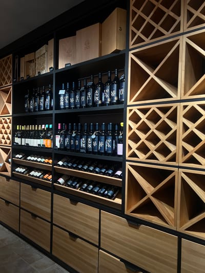 An interior view depicts a modern wine storage and display area, likely a retail space or cellar, located in Vinogradnoe, Russia. The scene is dominated by floor-to-ceiling shelving units crafted from light-colored wood and black metal frames.
On the left and right, extensive wooden wine racks feature a prominent diagonal grid pattern, forming numerous square and triangular compartments. These racks are largely empty in the upper sections but contain some horizontally stored bottles in the lower parts.
The central section comprises multiple black shelves displaying an organized collection of wine bottles. The top two shelves prominently feature dark-colored bottles, predominantly with black labels bearing white or light-colored script and the "УСАДЬБА МАРКОТ" (Usadba Markot) logo. Price tags are visible on many bottles (e.g., 5500, 3600, 2650, 3100 Rubles). Above these bottles, light-colored wooden gift boxes are stacked, some closed and some open, revealing straw packing material, also bearing the "УСАДЬБА МАРКОТ" emblem.
The lower central shelves present a mix of wine bottles, including some with lighter-colored labels (white, pink, light green, orange) standing upright. Below these, additional rows of dark-labeled bottles are laid horizontally in wooden cradles.
The entire lower portion of the shelving system consists of flush, light-colored wooden drawers with minimalist black horizontal handles. The floor is covered with large, square, light brown or greyish tiles. No individuals are present in the scene, and no active interactions are depicted. The lighting is focused, creating some shadowing while highlighting the displayed merchandise.