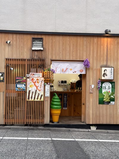 This is a street-level, full-frame photograph of a Japanese storefront, specifically a food and beverage establishment, in Kyoto, Japan. The scene depicts the facade of a building predominantly clad in light-colored vertical wooden planks. Above the wooden facade, the upper portion of the building presents a textured, off-white or light gray stucco wall, separated by a thin, dark horizontal band. On the far left of the wooden facade, a white security camera is mounted near the top. Below it, a vertical wooden slatted panel, appearing to be a partially open shutter or decorative screen, features a silver spotlight directed downwards. Adjacent to this, a cluster of posters is affixed to the wooden wall. These include a partially visible "ALCOHOL DRINK" sign, a poster advertising "BIG あわび" (abalone) with pricing, and another showcasing "特上牛串" (premium beef skewers). The most prominent poster in this section displays various skewers such as shrimp, scallops ("ホタテ串"), different cuts of meat, and cheese, alongside text mentioning "和牛コロッケ" (Wagyu Beef Croquette) for ¥300 and "神戶牛" (Kobe Beef). Towards the center, an open doorway reveals a counter inside the establishment. A person, partially visible from the back and wearing a dark cap, is behind the counter. A white 'noren' curtain, decorated with simple red illustrations of food items like dango (sweet dumplings) and onigiri (rice balls), hangs above the entrance. Clusters of artificial purple wisteria flowers adorn both sides of the 'noren'. To the right of the entrance, the wooden facade continues. A framed sign features a stylized black-and-white illustration of a Japanese woman and the text "そうすけ SOSUKE Japanese sake & food". Below this, a large, vibrant green poster advertises "MATCHA JAPAN traditional tea 抹茶 ソフトクリーム" (Matcha Soft Cream), showcasing a photorealistic image of green soft-serve ice cream in a black cup, and explicitly mentioning "宇治抹茶" (Uji Matcha), a renowned type of matcha from Kyoto Prefecture. Another cluster of artificial purple wisteria flowers hangs near this poster. A dark, modern outdoor light fixture is mounted on the upper right wooden wall, casting a warm yellow light. A large, three-dimensional statue of a green soft-serve ice cream cone, with a yellow waffle cone base marked with Japanese text ("抹茶のコーン"), stands on the sidewalk in front of the shop entrance, to the left of the doorway. It serves as a prominent visual advertisement. The ground consists of a gray asphalt sidewalk with a distinct white painted line running horizontally