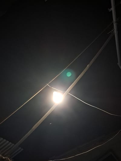 The image shows an outdoor nighttime scene in Vypasne, Ukraine. A single lightbulb, illuminated and emitting a bright white light, hangs from wires. These wires are suspended between utility poles and other supports, crisscrossing the dark sky. One of the wires runs diagonally across the foreground, passing close to the lightbulb. To the left and bottom of the image, a corrugated metal roof is visible, suggesting the presence of a structure beneath. The sky is dark, indicating nighttime, and a faint green lens flare is present in the upper left quadrant of the image, caused by the bright light source. There are no people or animals visible.