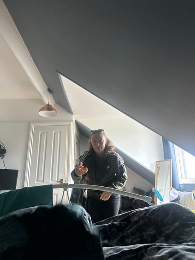 This image depicts a residential bedroom located within Salisbury, United Kingdom. The perspective is low, looking upwards from a bed in the foreground.
In the mid-ground, a young adult female with light brown hair, pulled back with sunglasses perched on her head, stands facing slightly left of the camera. She is wearing a dark olive green Adidas zip-up hoodie with three white stripes on the sleeves and dark trousers. Her left hand is raised, palm partially open, with light-colored nail polish visible, suggesting a gesture of speech or direction.
The room features a distinctive sloped attic ceiling, painted in two tones: the lower sections are white, transitioning to a dark grey or charcoal on the upper, angled planes. A copper-colored pendant light fixture hangs from the white section of the ceiling. To the left, a white panel door with a brass-colored handle is visible. Further left, a dark television screen rests on a surface. To the right, a window allows bright natural light into the room, with blue curtains partially visible. Beneath the window, a rectangular, wooden-framed mirror leans against the wall, and various items are present on a surface, including a radiator. The foreground is occupied by a bed covered with a dark, patterned duvet or blanket, with a white metal headboard (or footboard) partially obscured, draped with a dark green fabric.