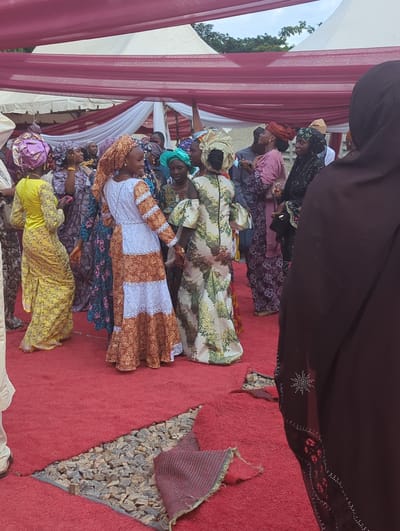 This image captures an outdoor social gathering in Kabala, Nigeria. The scene is illuminated by natural light under a partly cloudy sky, with white tents and draped maroon and white fabric forming an overhead canopy. The ground is covered with a bright red carpet, which has a triangular section cut out or missing in the foreground, revealing a bed of grey and light brown gravel. A crumpled piece of red, patterned fabric rests on the gravel.
Numerous individuals, predominantly women, are present, dressed in vibrant, patterned traditional African attire, including long dresses and elaborate headwraps (gele). In the center of the frame, two women appear to be interacting or dancing: one in a long yellow gown with a purple headwrap, and another in a white and orange patterned dress with an orange headwrap, facing each other. To their right, another woman wears a light-colored floral dress and matching headwrap. Other individuals, including men partially obscured, are visible in the midground and background, standing and conversing. In the immediate right foreground, the back and side of a person wearing a dark brown garment with intricate rhinestone or beaded embellishments on the sleeve are visible. The overall impression is a festive communal event.
