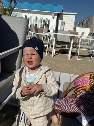 A baby wearing a dark blue cap and a light brown linen outfit sits in a gray chair on a beachside patio. The patio has white tables and chairs, and is attached to a building with a blue awning. Behind the building, a taller building is visible. A striped bag with a brown leather strap sits on another chair near the baby. The location is Middelkerke, Belgium.