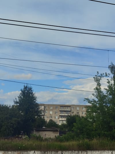 The image depicts a residential apartment building in the city of Фруктовая, Russia. The building is a multi-story concrete structure with a grid of windows and balconies. It is surrounded by lush green trees and vegetation. In the foreground, there is a low brick structure with a dark roof, possibly a shed or outbuilding. The sky is blue with scattered white clouds. Overhead, several black power lines traverse the frame horizontally. The overall scene suggests a typical urban or suburban residential area.