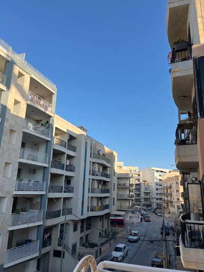 The image depicts a street scene in Saint Paul's Bay, Malta, under a clear blue sky. The view is dominated by apartment buildings with multiple balconies, typical of the urban architecture in the area. The buildings are primarily light-colored, with some featuring blue accents. Balconies are equipped with white or dark railings and some contain potted plants and laundry. Several cars are parked on the street and visible in motion, including a white hatchback, a dark sedan, and a red bus. People are sparsely visible, with a few individuals appearing on balconies or walking on the street. The environment suggests a residential urban setting, with the architecture and the presence of parked vehicles indicating it's likely a populated area. The sunlight casts shadows on the buildings, suggesting it's either morning or late afternoon.