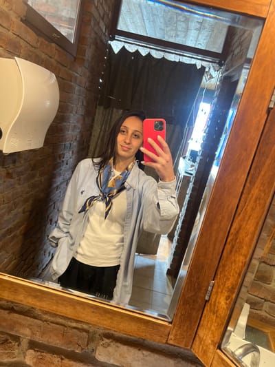 A female individual is depicted in a mirror selfie taken indoors within the city of Argveta, Georgia. The setting features exposed red brick walls and a prominent light-brown wooden-framed mirror. The individual, possessing dark, shoulder-length hair, is shown with a faint smile, holding a bright red smartphone (appearing to be an iPhone) in her right hand to capture the image. She is attired in a white t-shirt, an open light blue long-sleeved button-down shirt featuring a discernible polo player logo on the left chest, and a blue and yellow chain-link patterned scarf tied around her neck. Black lower garments are visible below the shirt. To the left of her reflection, a white wall-mounted paper towel dispenser is affixed to the brick wall. The mirror's upper portion reflects overhead windows exhibiting light refractions. The reflected background includes a dark, possibly curtained, doorway and a section of tiled flooring. A partial view of a white sink or counter edge is faintly visible in the lower right of the mirror's reflection.