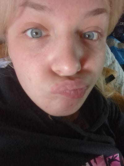 A close-up, chest-up selfie depicts a light-skinned female with blonde hair and blue eyes, looking directly at the camera with her lips puckered. Her face shows some freckling/blemishes. She wears a dark, possibly black, hooded sweatshirt with a partial pink and white graphic visible on the lower right. The background is blurred, showing light-colored fabric, likely bedding, and a brown object on the right, indicating an indoor, personal setting. The scene is located in the city of Area D (Elaho/Garibaldi), Canada.