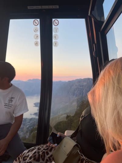 Inside a gondola car, two people are visible as the sun sets over a mountainous landscape with a body of water below. The person in the foreground is wearing a white t-shirt and looking down, their dark hair visible. Behind them, a person with blonde hair is partially visible, wearing a black top and a leopard-print shirt. They are holding a smartphone in their right hand. A light green purse is slung over their shoulder.
The gondola has large windows and a sunroof, offering panoramic views. Several circular icons are visible on the inside of the gondola windows, including a "no smoking" symbol. The sky shows a gradient of colors from pale blue to orange and pink, indicating sunset. The mountainous terrain is rugged and green, with buildings visible in the distance near the water. The location appears to be Škaljari, Montenegro. The overall mood is peaceful, with the sunset creating a serene atmosphere. There is no discernible text in the image.