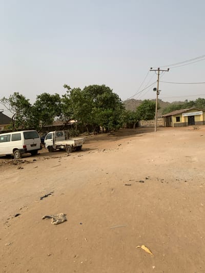 This image depicts an unpaved, dusty road scene in Bwari, Nigeria, under a pale, hazy sky. The foreground and mid-ground consist of reddish-brown dirt, with scattered refuse including a light-colored fabric item, a yellow leaf, and a blue plastic cap. On the left, a white passenger van is parked, followed by a smaller white pickup truck behind it. A cluster of green-leafed trees and the upper portion of a building's roof are visible on the far left. To the right, a single-story building with yellow walls and a brown base displays a sign reading 'THE LORD'S CHOSEN'. A wooden utility pole, equipped with multiple overhead wires and a loudspeaker, stands near the center-right, positioned in front of this building. The background reveals rocky outcrops and additional green trees. No individuals are visible, and no discernible actions or interactions are occurring within the frame.