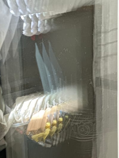 The image captures a distorted and blurry view of a display behind a glass surface. In the foreground, a dark container holds several items, including what appears to be a tan-colored object with yellow tassels or fringe. Behind this, several white fabric-like items are stacked, catching bright light.
Further back, a faint, stylized drawing of a cityscape with tall buildings and what seem to be figures walking on stairs is visible, rendered in thin white lines. Reflections and distortions from the glass create overlapping images and hazy effects. The background appears to be a dark, textured surface, possibly a wall or fabric.
The image does not provide any visual information to determine its location in the city of Trieste, Italy.