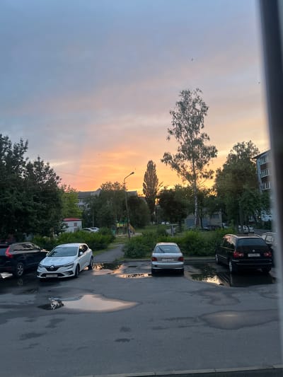 The image depicts an outdoor urban scene in Mazyr, Belarus, during a sunset. A paved parking lot with several cars is in the foreground. To the left, a black Volvo SUV is parked. In the center, a white Renault hatchback is parked, and to its right, a silver Hyundai sedan is visible. Further to the right, a black minivan is parked. Puddles of water are scattered across the asphalt, reflecting the colorful sky.
Beyond the parking lot, a green grassy area is dotted with trees and bushes. A children's playground with a slide and swings is situated in the middle ground. Apartment buildings and other structures are visible in the background, interspersed with more trees. The sky is a prominent feature, displaying a gradient of soft blue transitioning into vibrant orange and pink hues, indicative of a sunset. Several small birds are present in the sky. A street lamp stands tall near the playground. The overall impression is a quiet, residential area during the evening.