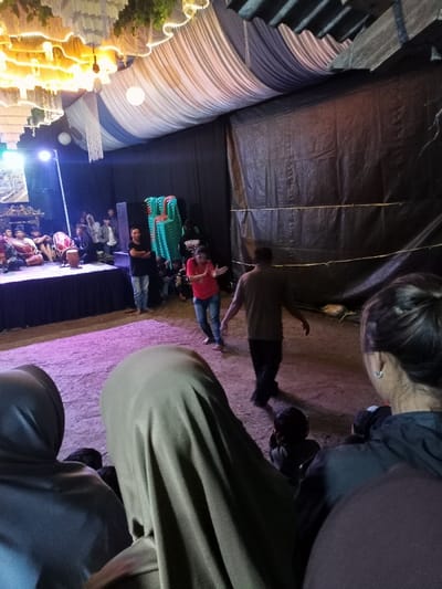 This image depicts a vibrant cultural performance or event held at night in Pangalengan, Indonesia, likely under a large tent or temporary structure. The setting features an elaborately decorated ceiling with white and blue draped fabric, adorned with festive string lights and hanging white globes, creating a warm, inviting glow.
In the foreground, several audience members are seated on the ground, observed from behind. Two individuals wear hijabs, one in a light olive green and another in a darker tone. Another person on the far right has their back to the camera, revealing a silver earring. A child's head is also visible among the seated audience members.
The middle ground showcases the primary activity. Two men are central to the scene: one in a red shirt and blue jeans is actively dancing or gesturing with his arms raised, while another in a brown t-shirt walks across the dirt ground, which is illuminated by a soft purple light. Behind them, stacked green plastic chairs are visible against a dark, possibly tarpaulin, wall.
To the left, a raised stage is brightly lit by a spotlight. On the stage, several performers are seated around traditional Indonesian musical instruments, specifically kendang (drums), indicating a musical performance. A light-colored banner or poster is displayed on the stage, though its text is unreadable due to glare. Another man stands near the stage, observing the performance. The background consists of dark tarpaulin walls, enclosing the event space. The overall impression is of a lively, community-focused gathering centered around music and dance.