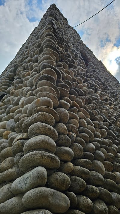 The image shows a low-angle, upward-looking view of a tall, triangular structure constructed entirely of smooth, rounded stones. The stones are of varying sizes and shades of gray and brown, and are meticulously stacked to form the sides of the structure. The perspective emphasizes the height and scale of the construction, with the peak disappearing into a cloudy sky. A thin black wire or cable is visible stretching diagonally across the upper right portion of the frame, against the sky. The overall impression is one of a unique architectural or artistic creation, likely an outdoor sculpture or building element. The image does not depict any people, specific actions, or interactions. The location is identified as the city of Krasnyy Kurgan, Russia, implying this structure is situated within an urban or semi-urban environment.