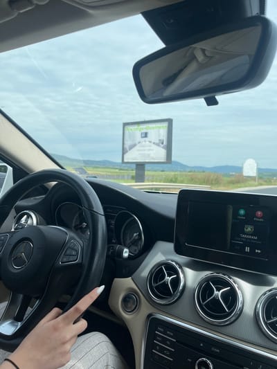 The image shows a view from inside a car, with a dashboard and steering wheel prominently featured. The steering wheel has a Mercedes-Benz logo. A hand with long white nails is visible, near the steering wheel. The car's infotainment system is displaying the song "TARARAM" by Emil TRF, DJ Damyan. Outside the car, a billboard advertising "Prestige Business" is visible. The landscape in the background consists of green fields and distant hills, indicating a rural or semi-rural area. The road is clearly visible. The text "Plovdiv" is seen on the car's display screen, which indicates that this photograph was likely taken in Plovdiv, Bulgaria. Given the billboard’s advertisement and the song selection, the overall setting hints at a blend of business and personal activity while driving in a semi-rural area outside of Plovdiv, Bulgaria. I cannot precisely determine the location as the city of Barakovo is not visible.