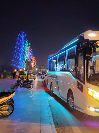 This nighttime image depicts a lively urban scene in Sa Pa, Vietnam. A large, white passenger bus, illuminated by its own bright headlights and prominent blue LED strip lighting along its side windows and roofline, dominates the right foreground, parked parallel to a paved sidewalk. Its interior shows light-colored seats and subtle decorative elements.
On the left, a row of large, abstractly-shaped trees, intricately decorated with vibrant blue, green, and red LED lights, stretches into the distance, providing significant ambient illumination. Several motorcycles are parked on the sidewalk: a dark blue or black motorcycle is in the midground, and the front wheel and partial body of a white and silver motorcycle are in the immediate left foreground.
In the midground, between the parked motorcycles and the illuminated trees, multiple individuals are visible on the sidewalk, some standing and others walking. One person in a light-colored jacket is clearly visible near a streetlamp which features a "WC" sign. Further down the street, additional vehicles, including cars and scooters, are parked. The environment consists of a dark night sky, a well-lit paved sidewalk, and a street curb, with reflections of the artificial lighting visible on the ground, suggesting a wet or reflective surface. Distant buildings and lights are faintly discernible in the background.