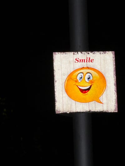 A wooden sign featuring a cartoon smiley face and the word "Smile" is mounted on a dark, cylindrical pole. The sign is rectangular with rounded corners and has a distressed, weathered appearance with visible wood grain and peeling paint along the edges. The smiley face is bright orange with large blue eyes, rosy cheeks, and a wide, open mouth showing white teeth and a red tongue. The word "Smile" is written in red, stylized lettering above the smiley face. The background of the image is predominantly black, suggesting it was taken at night or in a dimly lit environment. The pole is positioned slightly off-center to the right, and a hint of dark foliage is visible in the lower left corner. This image was taken in the city of Pravets, Bulgaria.